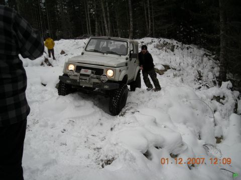 White Suzuki Samurai in the snow on a wooded trail with two men.
