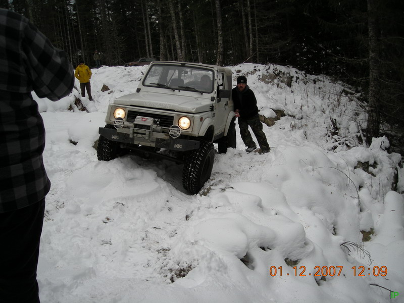 White Suzuki Samurai in the snow on a wooded trail with two men.