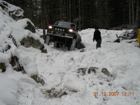 Dark blue Ford Bronco driving up a snowy, rocky hill in a forest.