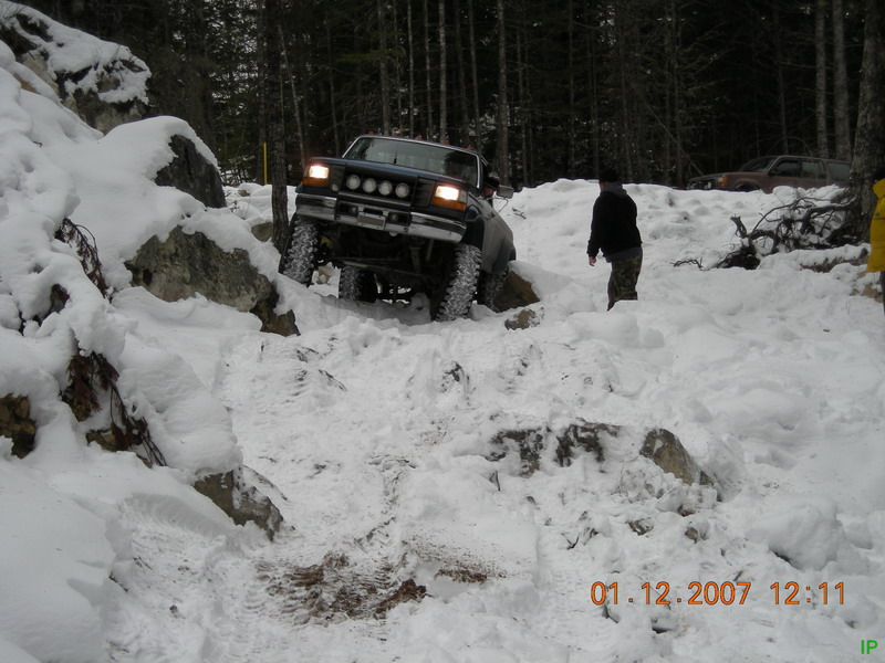 Dark blue Ford Bronco driving up a snowy, rocky hill in a forest.