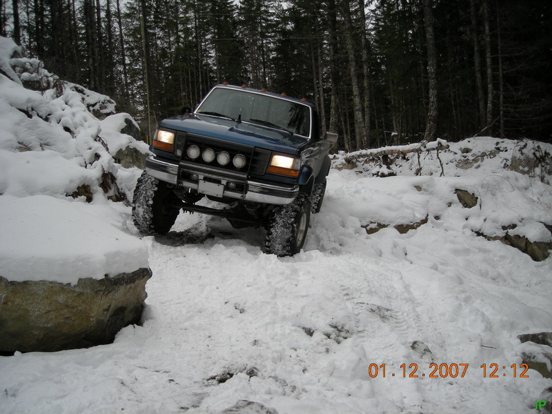 Lifted blue Ford truck driving on a snowy, rocky trail in a forest.