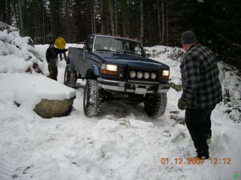 Lifted blue Ford pickup truck on a snowy trail with people.