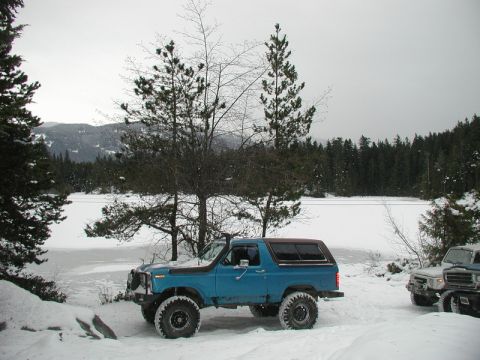 Blue Ford Bronco parked in the snow by a frozen lake with a forest and mountains.