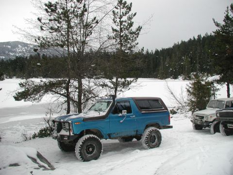 Blue Ford Bronco with large tires parked in snow near a frozen lake and forest.