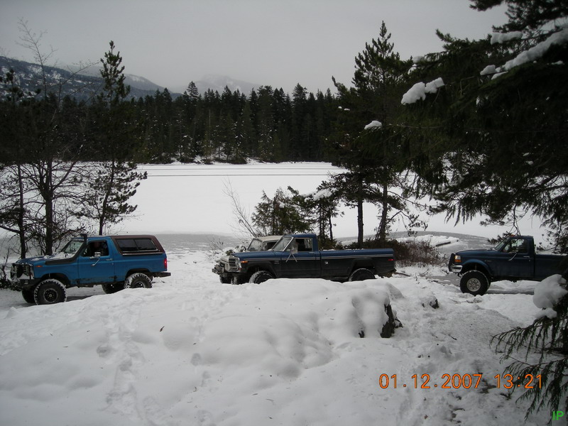 Three pickup trucks and a blue SUV in the snow by a frozen lake.