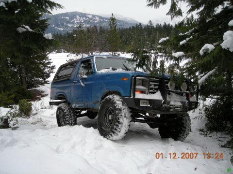 Blue 1985 Ford Bronco in snow with custom bumper and large tires.