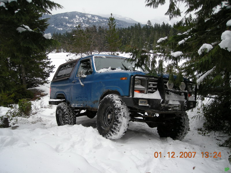 Blue 1985 Ford Bronco in snow with custom bumper and large tires.