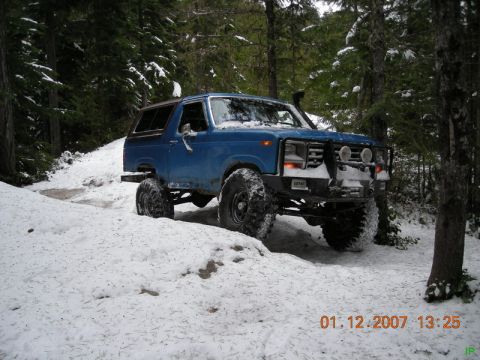 Blue 1985 Ford Bronco on a snowy forest trail with large tires.