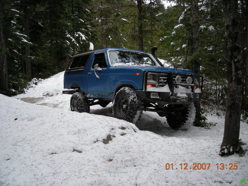 Blue 1985 Ford Bronco on a snowy forest trail with large tires.