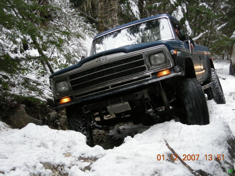 Dark blue Jeep truck off-roading in snow, front tire elevated over rocks.