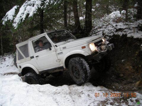 White Suzuki Samurai off-roading on a snowy trail in a forest.