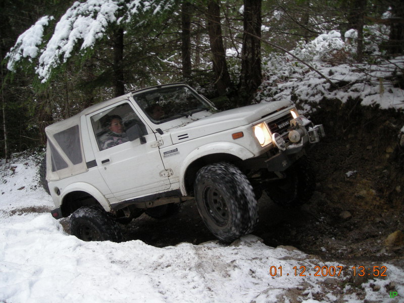 White Suzuki Samurai off-roading on a snowy trail in a forest.