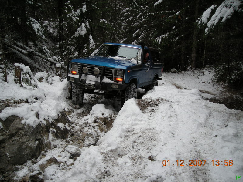 Blue 1980s Ford Bronco off-roading on a snowy forest trail.