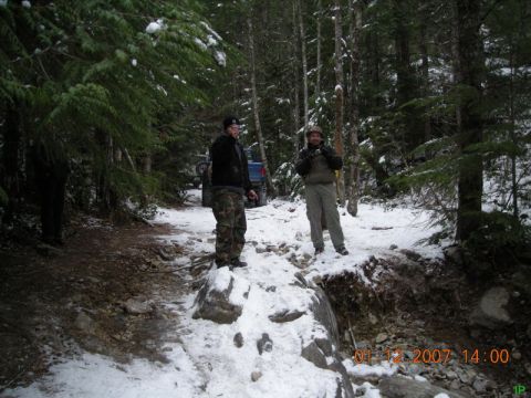 Two men on a snowy trail next to a blue Ford Bronco.