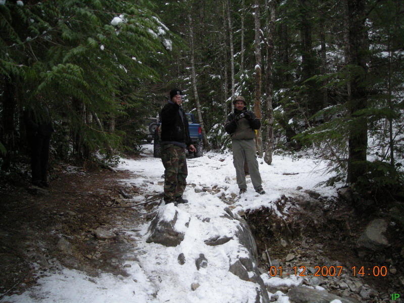 Two men on a snowy trail next to a blue Ford Bronco.