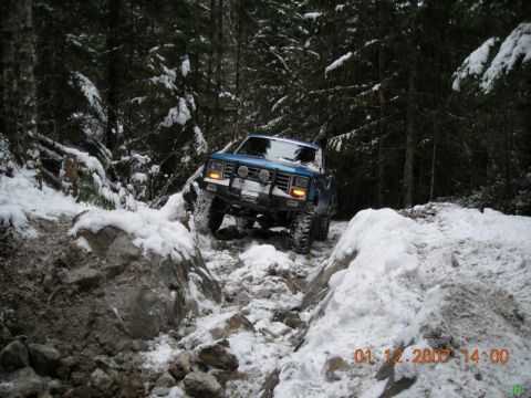 Blue 1980s Ford Bronco off-roading on a snowy, rocky trail in a forest.