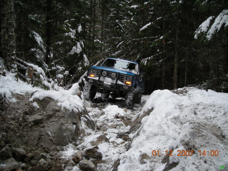 Blue 1980s Ford Bronco off-roading on a snowy, rocky trail in a forest.