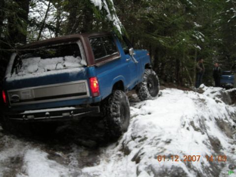 Blue 1980s Ford Bronco off-roading on a snowy trail.