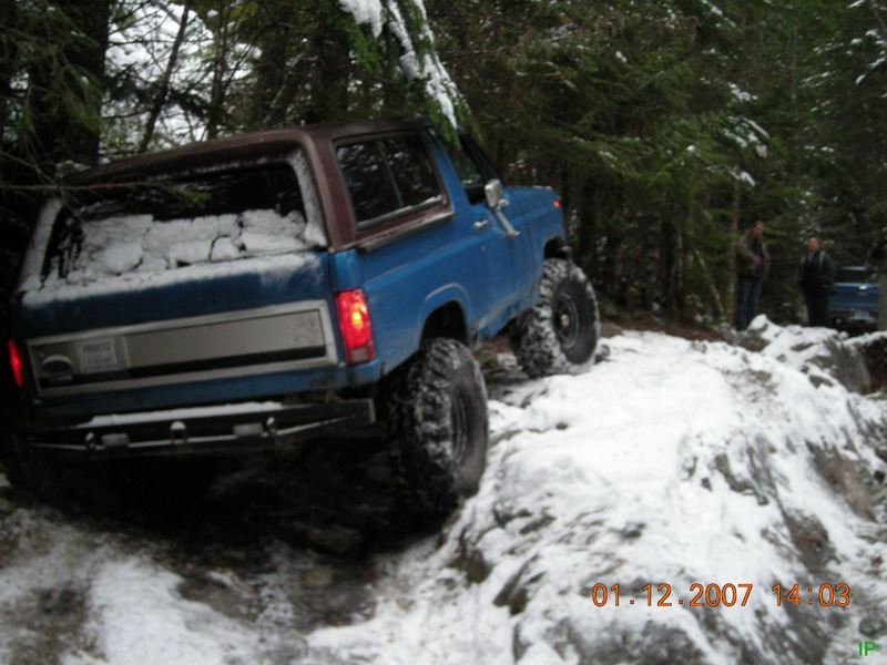 Blue 1980s Ford Bronco off-roading on a snowy trail.