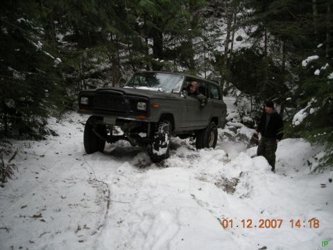 Lifted dark green Jeep Cherokee driving through snow in a forest with two men nearby.