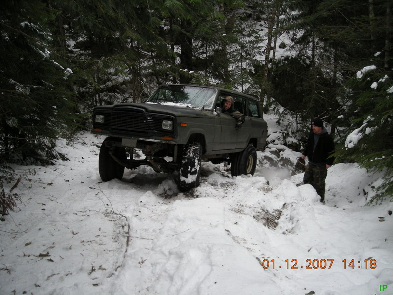 Lifted dark green Jeep Cherokee driving through snow in a forest with two men nearby.