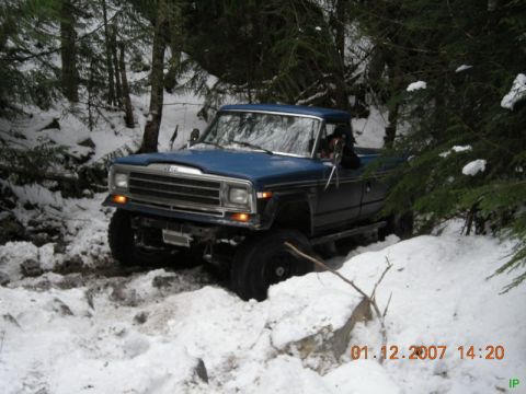 Blue Jeep pickup truck driving on a snowy, wooded trail.