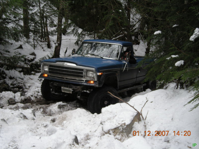 Blue Jeep pickup truck driving on a snowy, wooded trail.