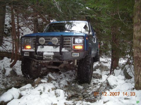 Blue 1985 Ford Bronco off-roading on a snowy trail with trees.