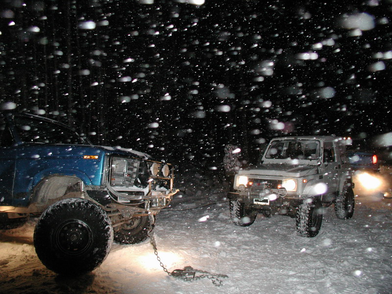 Blue Ford Bronco being towed by a silver SUV in a snowy forest at night.