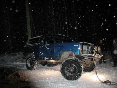 Lifted blue Ford Bronco in snow at night with people behind it.