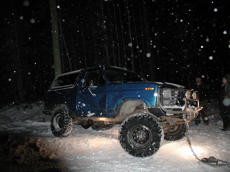 Lifted blue Ford Bronco in snow at night with people behind it.