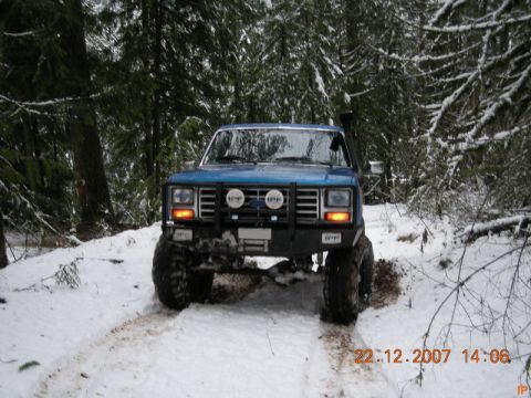 Blue Ford Bronco driving on a snowy forest road with large off-road tires and snorkel.
