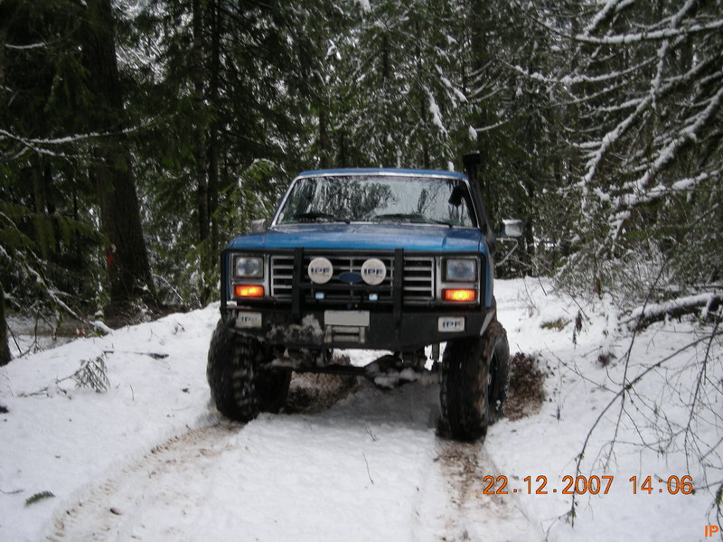Blue Ford Bronco driving on a snowy forest road with large off-road tires and snorkel.