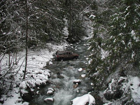 Brown SUV driving through a snowy stream in a forest.