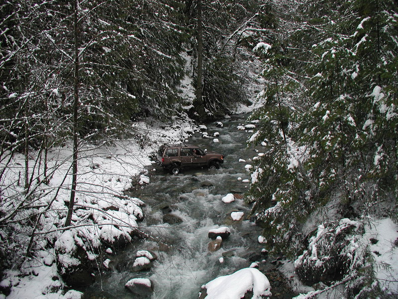 Brown SUV driving through a snowy stream in a forest.