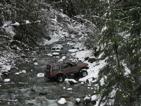 Lifted brown SUV driving through a rocky river in a snowy forest.