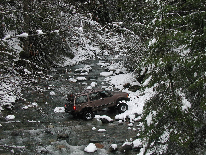 Lifted brown SUV driving through a rocky river in a snowy forest.