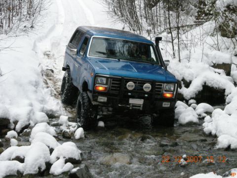 Blue Ford Bronco driving through a snowy stream with off-road tires and IPF lights.