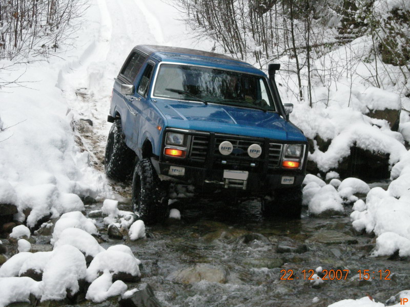 Blue Ford Bronco driving through a snowy stream with off-road tires and IPF lights.