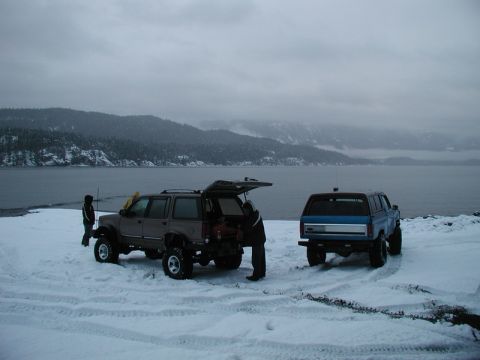 Two lifted SUVs on a snowy shore with a lake and mountains.