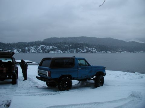 Blue Ford Bronco parked in snow by water with mountains in background.