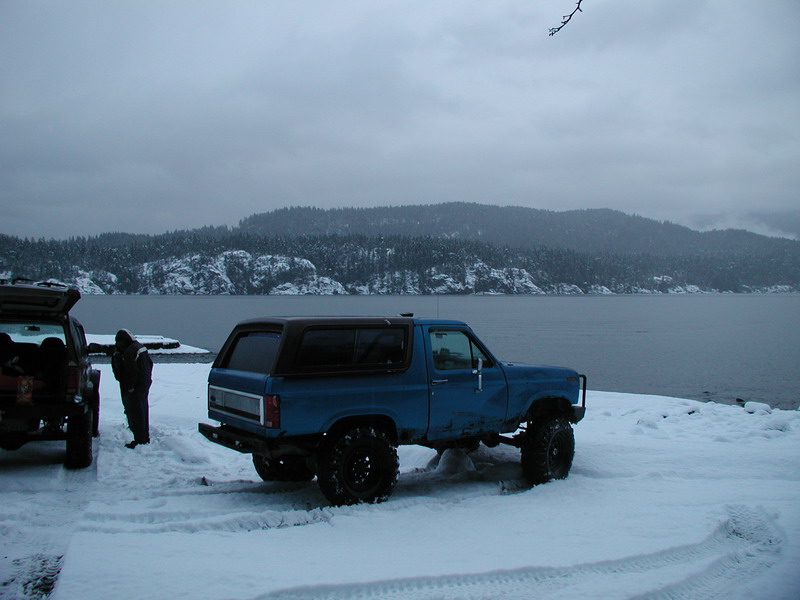 Blue Ford Bronco parked in snow by water with mountains in background.