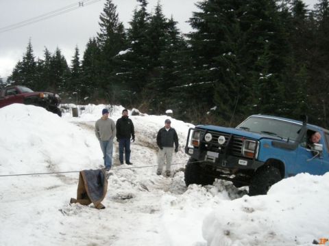Blue 1985 Ford Bronco stuck in snow being winched, with people around.