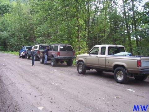 1985 Ford Bronco - A line of modified SUVs and a pickup truck are parked on a dirt road in a wooded area. The lead pickup truck is a tan Ford Ranger with off-…