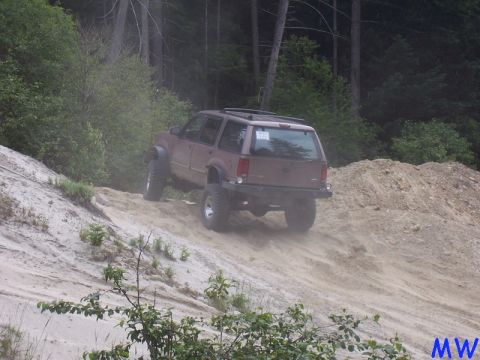 1985 Ford Bronco - A brown SUV is driving up a steep, sandy hill in a wooded area. The vehicle has large tires and appears to be modified for off-road use.
