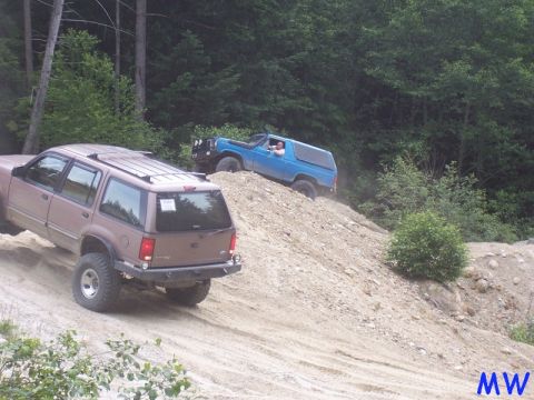 1985 Ford Bronco - A brown SUV is driving up a dirt hill, while a blue SUV is driving up the same hill behind it. The scene is set in a wooded area.