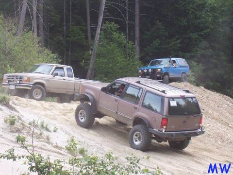 1985 Ford Bronco - Three off-road vehicles are driving up a sandy hill in a wooded area. A tan pickup truck is in the foreground, followed by a brown SUV, and…