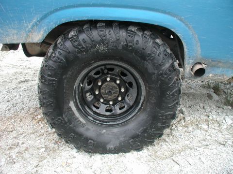 Close-up of a blue Ford Bronco's rear off-road tire and black steel wheel on a gravel surface.