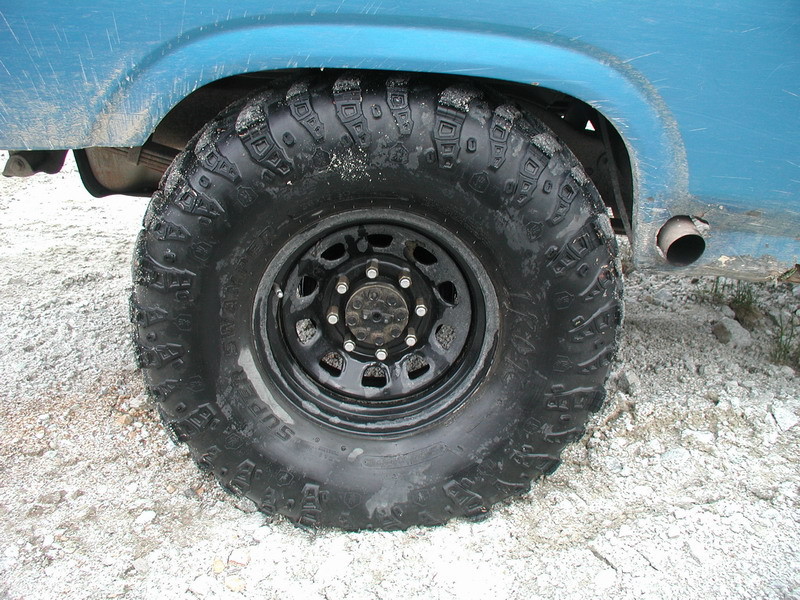 Close-up of a blue Ford Bronco's rear off-road tire and black steel wheel on a gravel surface.