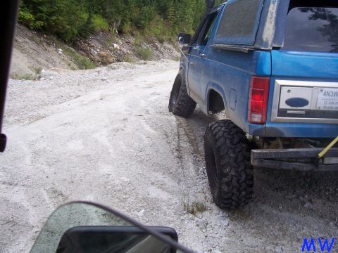 1985 Ford Bronco - The rear passenger side of a lifted blue Ford Bronco is shown on a gravel road with trees and a hillside in the background. The vehicle has…